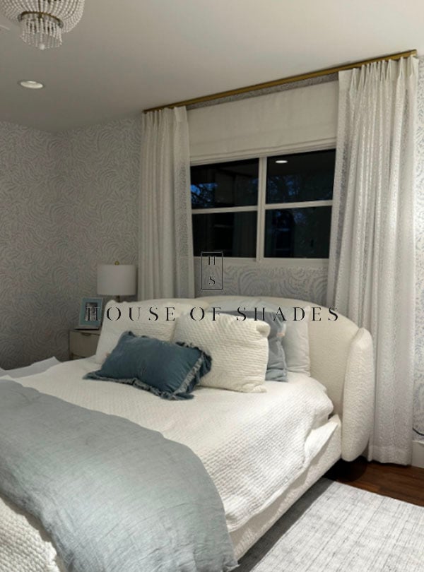 White pleated drapery framing a bedroom window with a Roman shade.